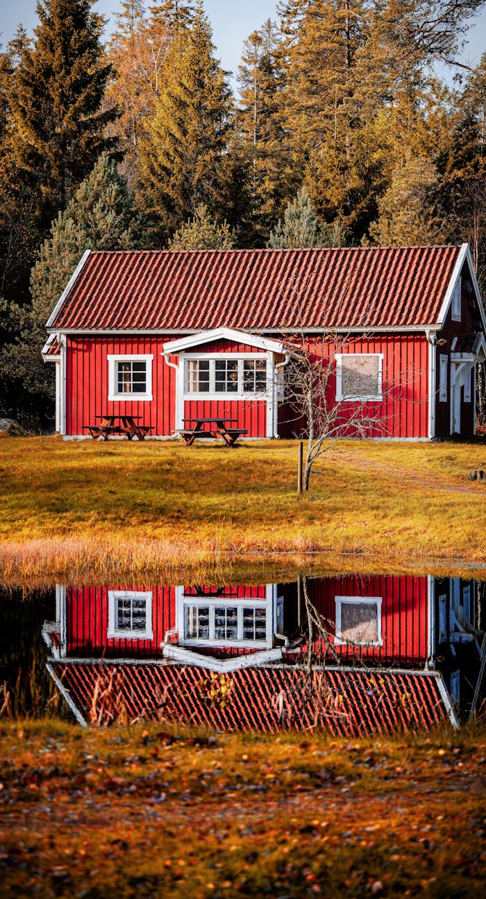 Red cabin reflected on a tranquil lake surrounded by autumn foliage, showcasing serene nature.