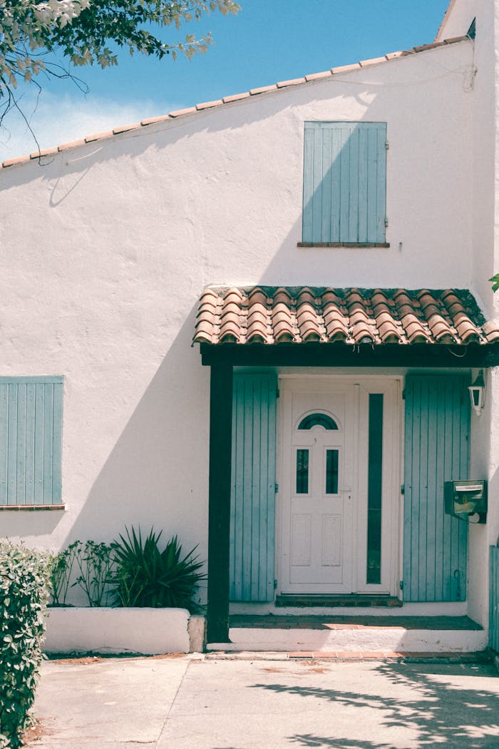 Exterior of white residential stone cottage with blue shutters and tiled canopy over entrance door on sunny weather